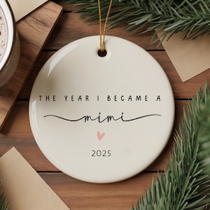 May include: A round, off-white ceramic ornament with gold string. The text "THE YEAR I BECAME A mimi" is written in black script, with a small pink heart and the year "2025" below. The ornament is on a wooden surface with greenery.
