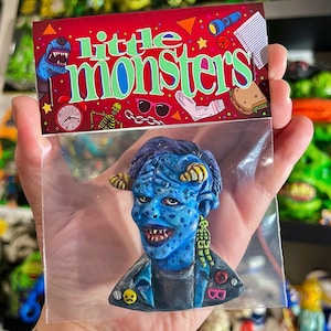 May include: A blue monster figurine with yellow horns and a black jacket is held in a clear plastic bag. The figurine has a detailed face and is part of the "Little Monsters" collection, as indicated by the red card with the text.