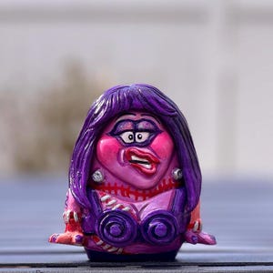 May include: A hand-painted figurine of a woman with purple hair and a dramatic expression. The figure has a pink face with exaggerated features, wearing a purple top with circular accents. The figure is on a blue surface.