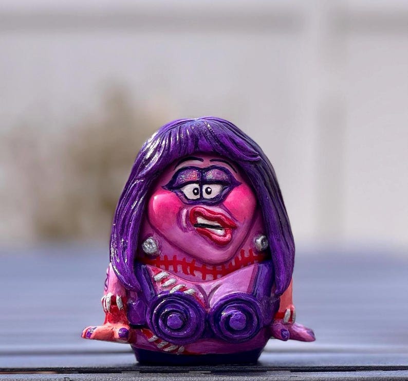 May include: A hand-painted figurine of a woman with purple hair and a dramatic expression. The figure has a pink face with exaggerated features, wearing a purple top with circular accents. The figure is on a blue surface.