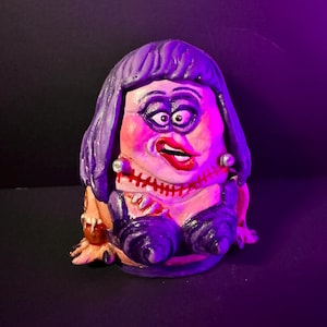 May include: A hand-painted ceramic figurine of a character with exaggerated features. The figure has purple hair, heavy makeup, and a stitched mouth. It wears a black outfit with a low neckline and has a grotesque, yet whimsical appearance.