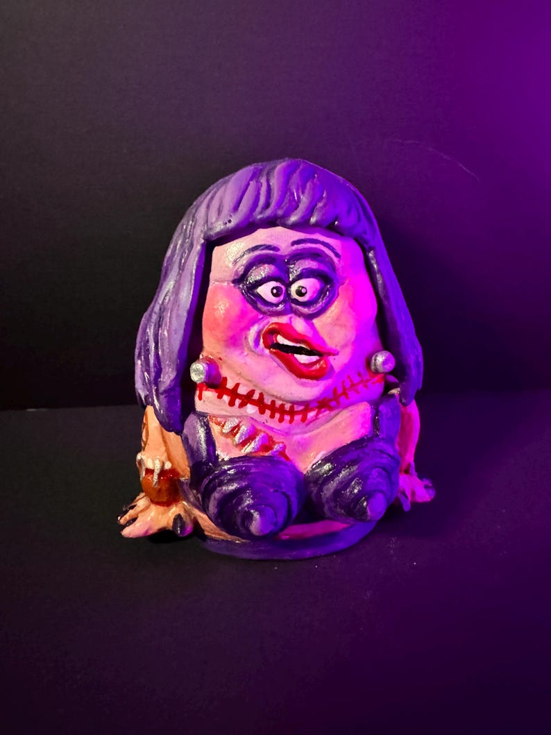 May include: A hand-painted ceramic figurine of a character with exaggerated features. The figure has purple hair, heavy makeup, and a stitched mouth. It wears a black outfit with a low neckline and has a grotesque, yet whimsical appearance.