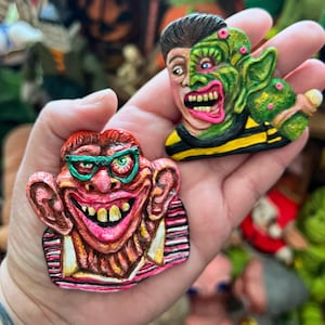 May include: Two grotesque, colorful, handmade face sculptures. One features a red-haired face with glasses and a wide grin. The other is a split face, half human and half green monster. Both are held in a hand, with other similar sculptures in the background.