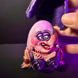 May include: A hand-painted, whimsical figurine of a character with an egg-shaped head, rouged cheeks, and a red lipstick mouth. The figure wears a purple outfit and has a matching purple wig. The figurine is a unique piece of art.