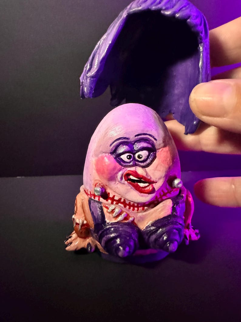 May include: A hand-painted, whimsical figurine of a character with an egg-shaped head, rouged cheeks, and a red lipstick mouth. The figure wears a purple outfit and has a matching purple wig. The figurine is a unique piece of art.