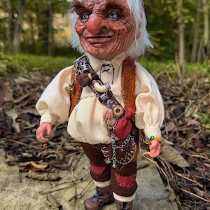 May include: A detailed doll of an elderly figure with a weathered face, blue eyes, and white hair. The doll wears a cream-colored shirt, brown pants, and boots. Accessories include a belt with pouches and keys, and a beaded bracelet. The background is a blurred forest.