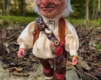 Könnte beinhalten: Detaillierte Puppe einer älteren Figur mit zerfurchtem Gesicht, blauen Augen und weißem Haar. Die Puppe trägt ein cremefarbenes Hemd, braune Hosen und Stiefel. Zu den Accessoires gehören ein Gürtel mit Beuteln und Schlüsseln sowie ein Perlenarmband. Der Hintergrund ist ein verschwommener Wald.