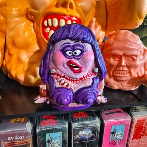 May include: A collection of colorful, grotesque monster figures, including a purple-haired, pink-faced character with a stitched neck and a large orange monster with a gaping mouth. VHS tapes are visible on a shelf below.