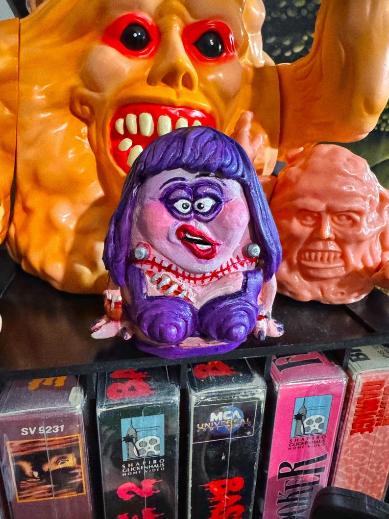 May include: A collection of colorful, grotesque monster figures, including a purple-haired, pink-faced character with a stitched neck and a large orange monster with a gaping mouth. VHS tapes are visible on a shelf below.