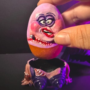 May include: A whimsical, hand-painted egg-shaped sculpture with a face, held above a matching base. The egg features a pink face with exaggerated makeup, a stitched mouth, and a cigar. The base is a miniature pair of blue pants.