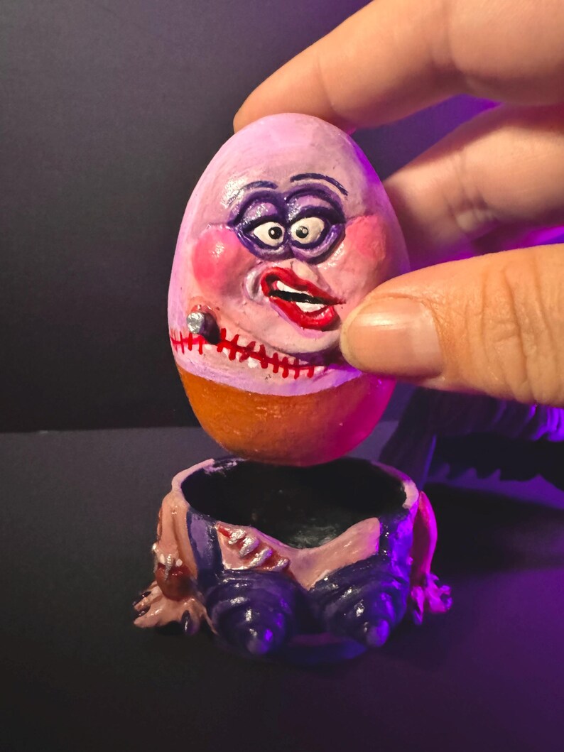 May include: A whimsical, hand-painted egg-shaped sculpture with a face, held above a matching base. The egg features a pink face with exaggerated makeup, a stitched mouth, and a cigar. The base is a miniature pair of blue pants.