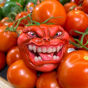 May include: A carved tomato with a grinning face, complete with teeth and a tongue, sits among a pile of fresh, ripe tomatoes. The carved tomato is a vibrant red color, with detailed facial features. Green tomatoes are visible in the background.