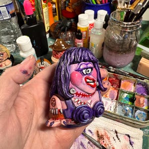 May include: A hand-painted, cartoon-style figurine with a purple dress, hair, and makeup. The figurine has a stitched mouth, large red lips, and a pearl necklace. The figure is holding a small, round object. The background includes art supplies.
