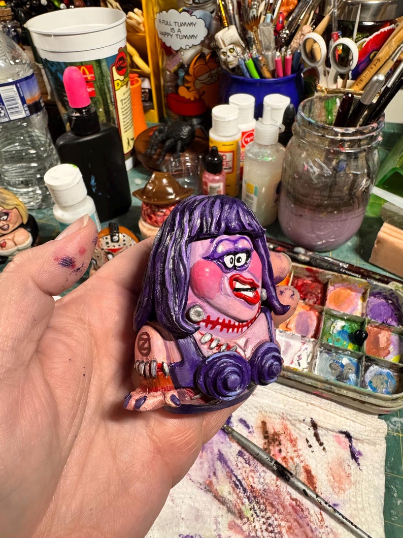 May include: A hand-painted, cartoon-style figurine with a purple dress, hair, and makeup. The figurine has a stitched mouth, large red lips, and a pearl necklace. The figure is holding a small, round object. The background includes art supplies.