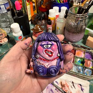 May include: A hand-painted, whimsical ceramic figurine of a woman with purple hair, a stitched neck, and a purple bustier. The figurine has exaggerated features and is held in a person's hand, with a colorful art studio background.