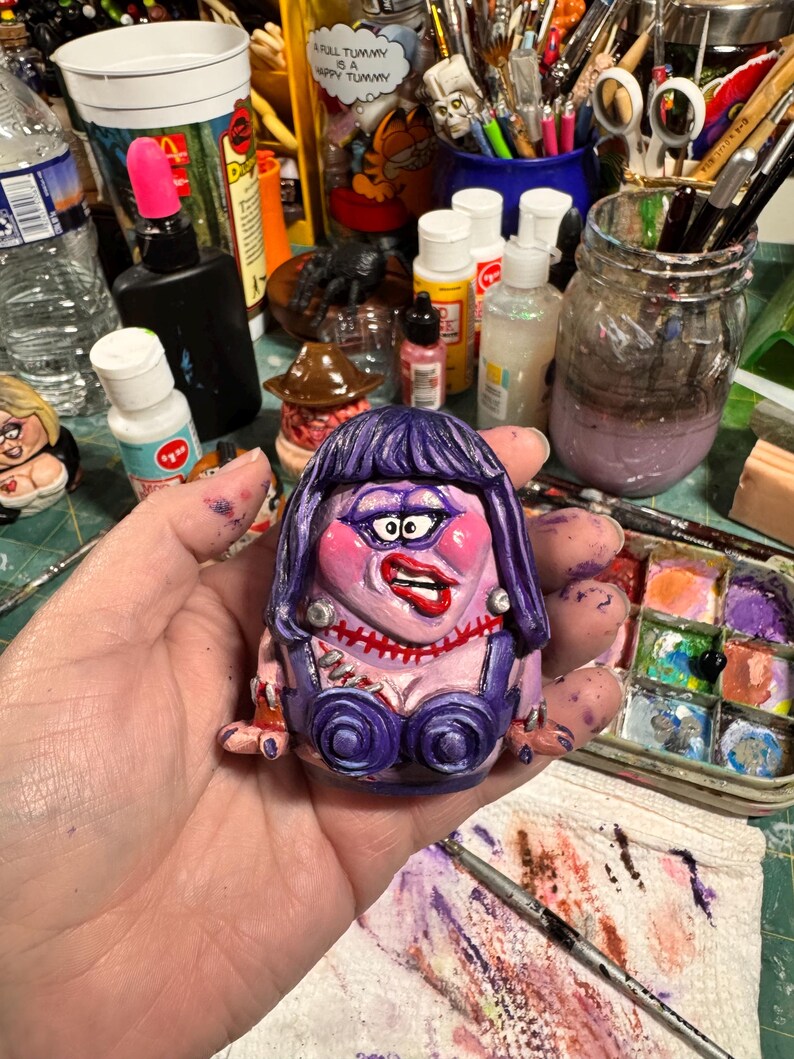 May include: A hand-painted, whimsical ceramic figurine of a woman with purple hair, a stitched neck, and a purple bustier. The figurine has exaggerated features and is held in a person's hand, with a colorful art studio background.