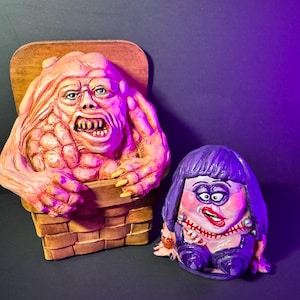 May include: Two grotesque, hand-painted sculptures. One is a fleshy, pinkish-orange creature emerging from a basket. The other is a purple-haired figure with a painted face, wearing a purple outfit. The background is dark with a purple light.