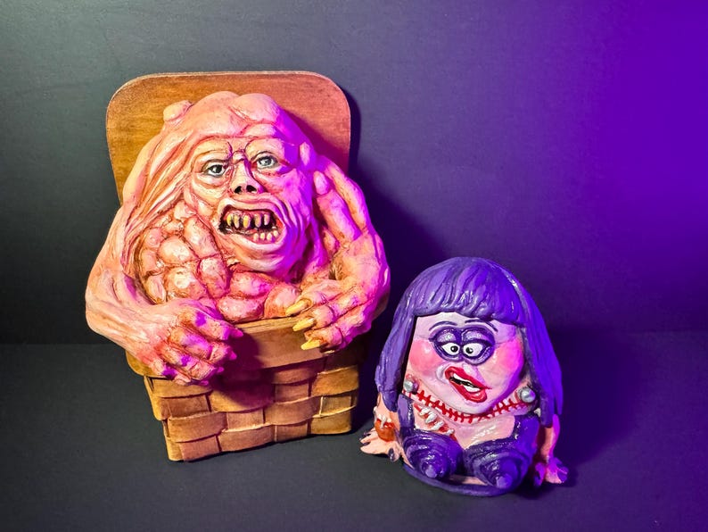 May include: Two grotesque, hand-painted sculptures. One is a fleshy, pinkish-orange creature emerging from a basket. The other is a purple-haired figure with a painted face, wearing a purple outfit. The background is dark with a purple light.