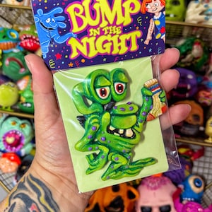 May include: A green monster figurine with purple spots and red eyes, holding a sock. The figurine is attached to a green card with the text "BUMP IN THE NIGHT" in yellow and blue. The background features other monster figurines.