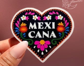May include: A heart-shaped sticker featuring the word "MEXICANA" in white. The sticker has a black background adorned with vibrant floral patterns in pink, orange, blue, and white. The sticker is held by a hand.