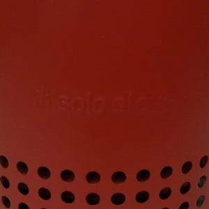 May include: A red metal Solo Stove with a series of evenly spaced circular holes near the base. The Solo Stove logo is embossed on the side. The stove is designed for outdoor use.