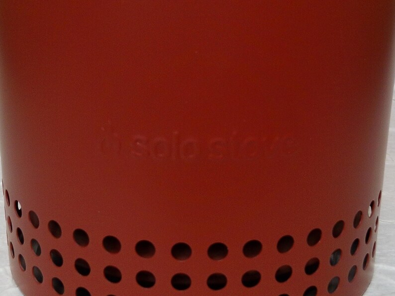 May include: A red metal Solo Stove with a series of evenly spaced circular holes near the base. The Solo Stove logo is embossed on the side. The stove is designed for outdoor use.