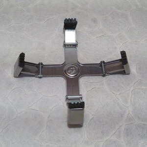 May include: A silver-coloured, cross-shaped tool with four adjustable black rubber grips. The tool has a central circular hub and is designed to hold an object in place. The grips are positioned at the ends of the cross arms.