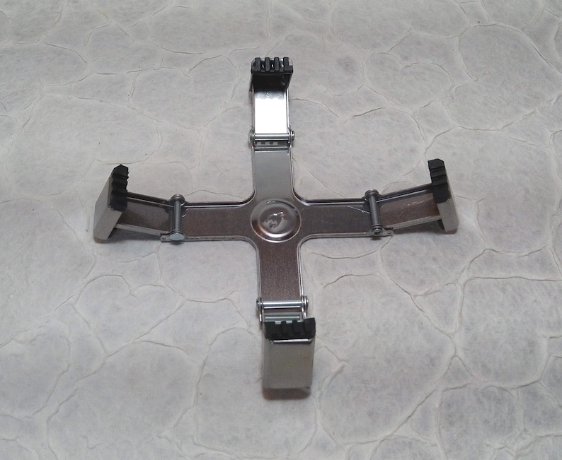 May include: A silver-coloured, cross-shaped tool with four adjustable black rubber grips. The tool has a central circular hub and is designed to hold an object in place. The grips are positioned at the ends of the cross arms.
