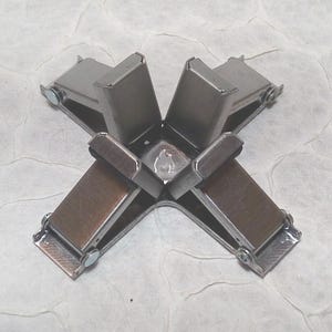May include: A metal corner clamp with four arms, each featuring a brown pad and a silver-coloured metal frame. The clamp is designed to hold corners together. The background is a textured, off-white surface.