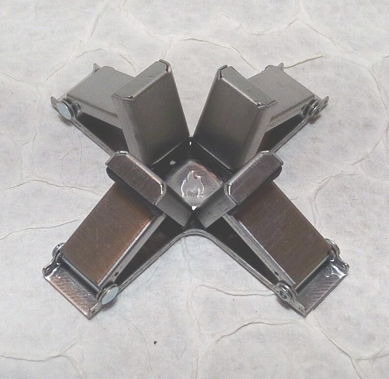 May include: A metal corner clamp with four arms, each featuring a brown pad and a silver-coloured metal frame. The clamp is designed to hold corners together. The background is a textured, off-white surface.