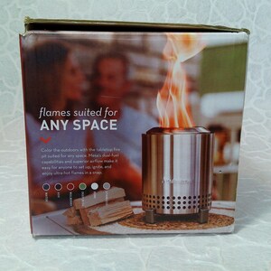 May include: A product box showing a stainless steel tabletop fire pit with flames. The box text reads "flames suited for ANY SPACE." The box also shows wood and color options. The fire pit is designed for easy setup and use.