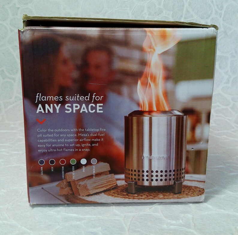May include: A product box showing a stainless steel tabletop fire pit with flames. The box text reads "flames suited for ANY SPACE." The box also shows wood and color options. The fire pit is designed for easy setup and use.