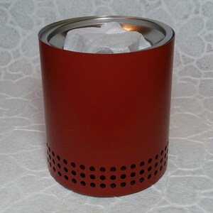 May include: A cylindrical, red metal container with a silver rim and small holes near the base. The interior contains white packing material. The container is designed for outdoor use. The height is approximately 15 cm.