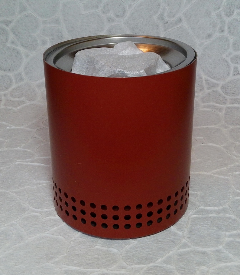 May include: A cylindrical, red metal container with a silver rim and small holes near the base. The interior contains white packing material. The container is designed for outdoor use. The height is approximately 15 cm.