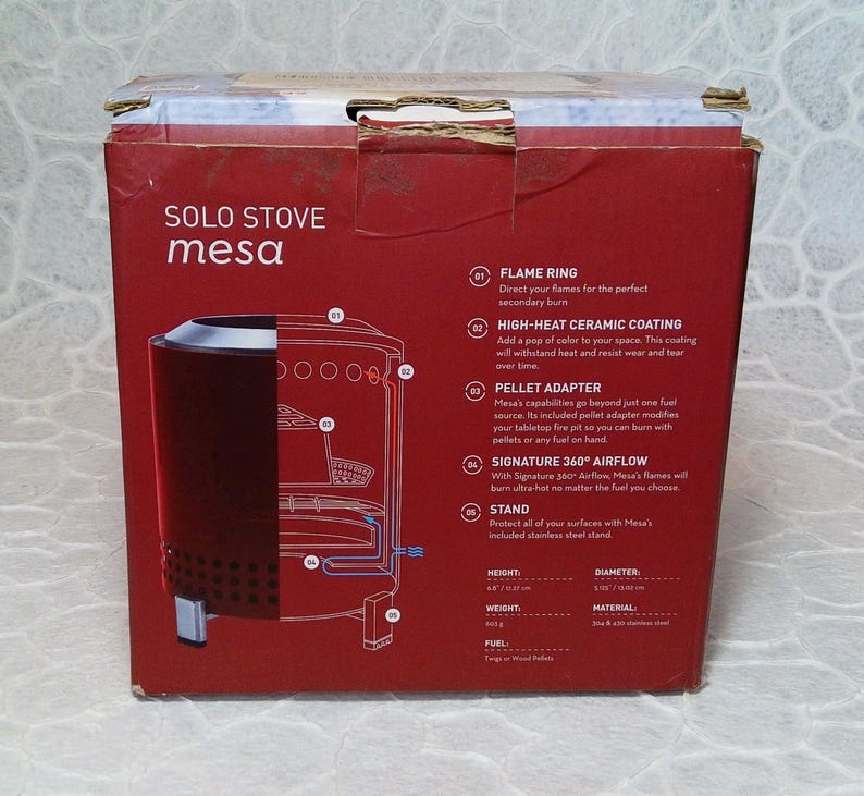 May include: A red box for a Solo Stove Mesa tabletop fire pit. The box features a diagram of the fire pit and lists features like a flame ring, high-heat ceramic coating, and pellet adapter. The box also lists the height as 17.27 cm and the diameter as 14.73 cm.