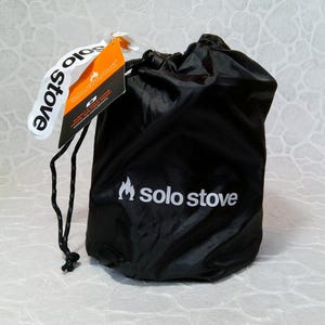 May include: A black drawstring bag with the words "solo stove" printed in white. A white tag with the words "solo stove" is attached to the bag. An orange and black tag is also attached to the bag.