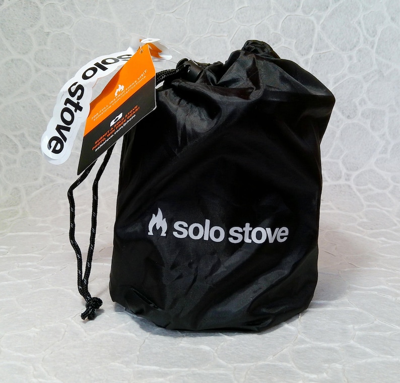 May include: A black drawstring bag with the words "solo stove" printed in white. A white tag with the words "solo stove" is attached to the bag. An orange and black tag is also attached to the bag.