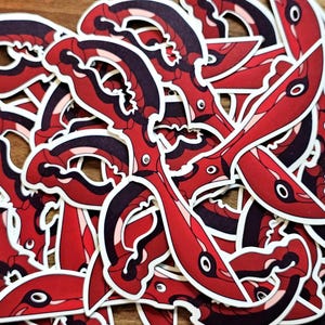 May include: A collection of red and black stickers with a stylized design. The stickers have a white border and feature a repeating pattern of curved shapes and eye-like details. The stickers are scattered on a wooden surface.