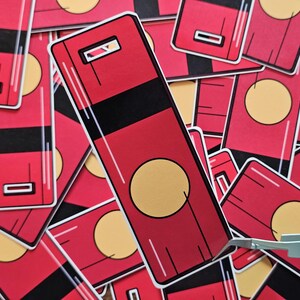 May include: Red and black rectangular stickers with a gold circle and a rectangular cutout. The stickers are arranged in a pile, with one being held by tweezers.