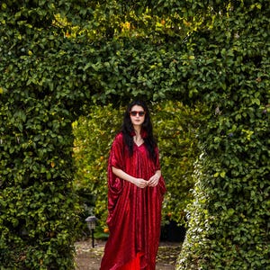 May include: A woman wearing a long, red sequined dress and matching red shoes stands in front of a green hedge archway. The dress has a V-neck and wide sleeves. She is also wearing red sunglasses.