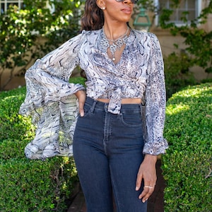 Peut inclure: Un chemisier portefeuille à imprimé peau de serpent gris et bleu marine, associé à un jean en denim foncé. Le chemisier a des manches longues évasées. Un collier argenté complète la tenue.