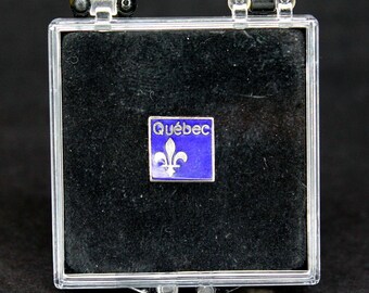 May include: A small, square pin featuring the word "Québec" and a fleur-de-lis symbol on a blue background. The pin is displayed inside a clear plastic box with a black velvet interior.