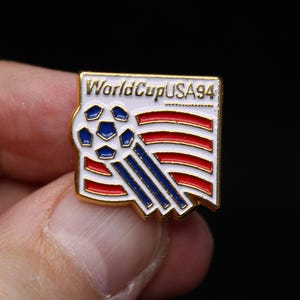 May include: A square-shaped metal enamel pin featuring the text WorldCupUSA94 in gold lettering. The design includes a white background with a blue and white soccer ball graphic and stylized red and blue stripes extending from the ball.