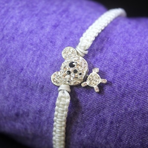 May include: White string bracelet with a silver charm featuring a koala bear with rhinestones and a smaller silver charm with rhinestones.
