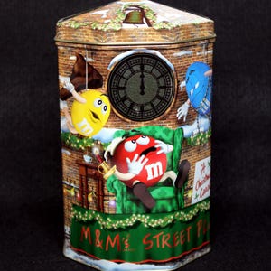 May include: A red, yellow, and blue M&M's Christmas tin with a clock tower design. The tin features a red M&M sitting in a green armchair, a yellow M&M hanging from a rope, and a blue M&M hanging from a chain. The tin says "M&M's Street Place" and "A Christmas Carol".