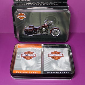 May include: Two decks of playing cards in a silver tin with a Harley-Davidson motorcycle graphic. The tin has the words "Motor Harley-Davidson Cycles" and "An American Legend".