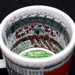 May include: White ceramic mug with a stadium scene illustration. The interior features a green field with the word "FURAC&Atilde;O" and a crowd of people with red and black balloons. The exterior has a red and gray design.
