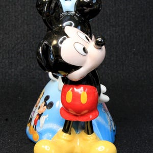 May include: A ceramic figurine of Mickey Mouse wearing red shorts with yellow buttons and black shoes. Mickey is standing with his head tilted to the side and his right hand on his chin. He is wearing a blue bow and is standing on a blue and white background with other Disney characters.