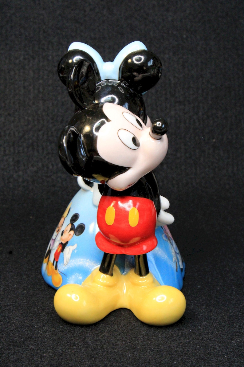 May include: A ceramic figurine of Mickey Mouse wearing red shorts with yellow buttons and black shoes. Mickey is standing with his head tilted to the side and his right hand on his chin. He is wearing a blue bow and is standing on a blue and white background with other Disney characters.