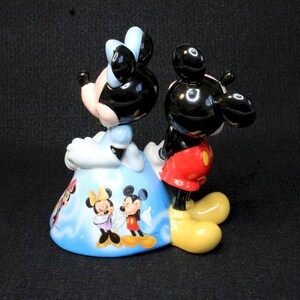 May include: A ceramic figurine of Mickey Mouse and Minnie Mouse. Mickey is standing behind Minnie, who is wearing a blue dress with a white and blue pattern. The figurine is painted in bright colors and features a whimsical design.